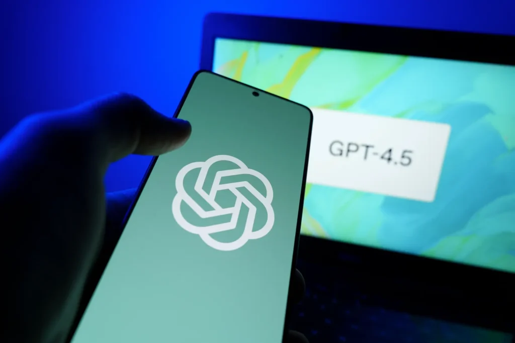 A hand holding a smartphone displaying the OpenAI logo, with a laptop in the background showing GPT-4.5 on-screen.