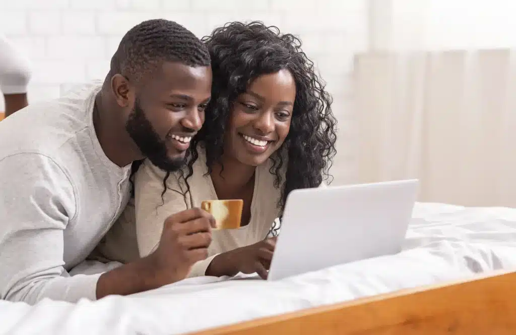 A smiling couple lying on a bed using a laptop and holding a credit card, symbolizing an online shopping experience enhanced by Hyper-personalization AI.