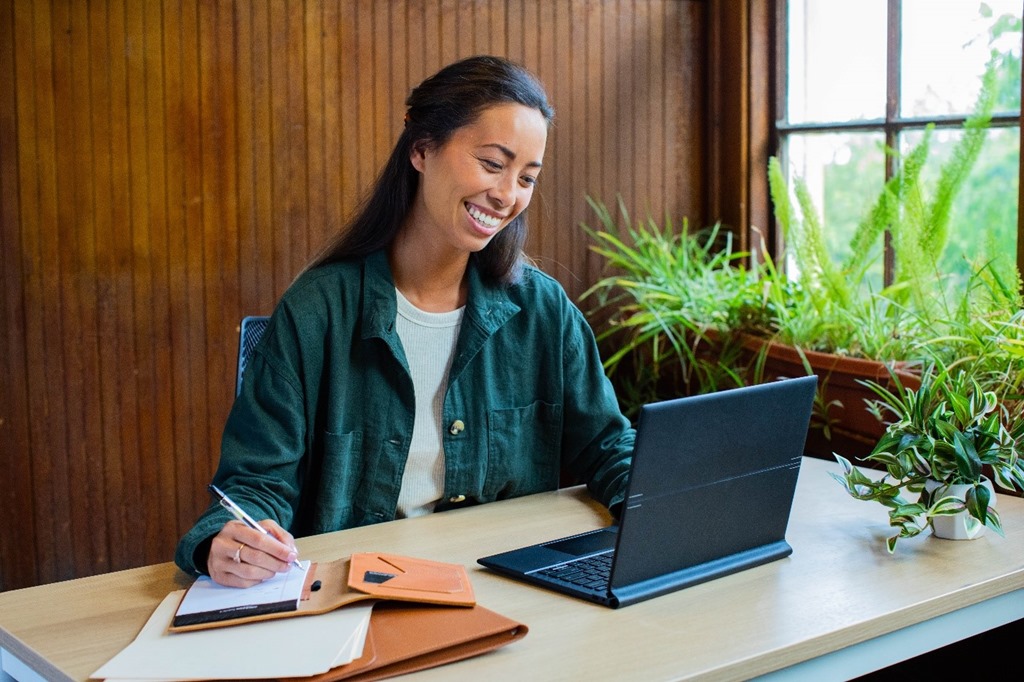 A woman sits at her desk and is smiling at her laptop in front of her. She holds a pen in her right hand and writes in a notebook