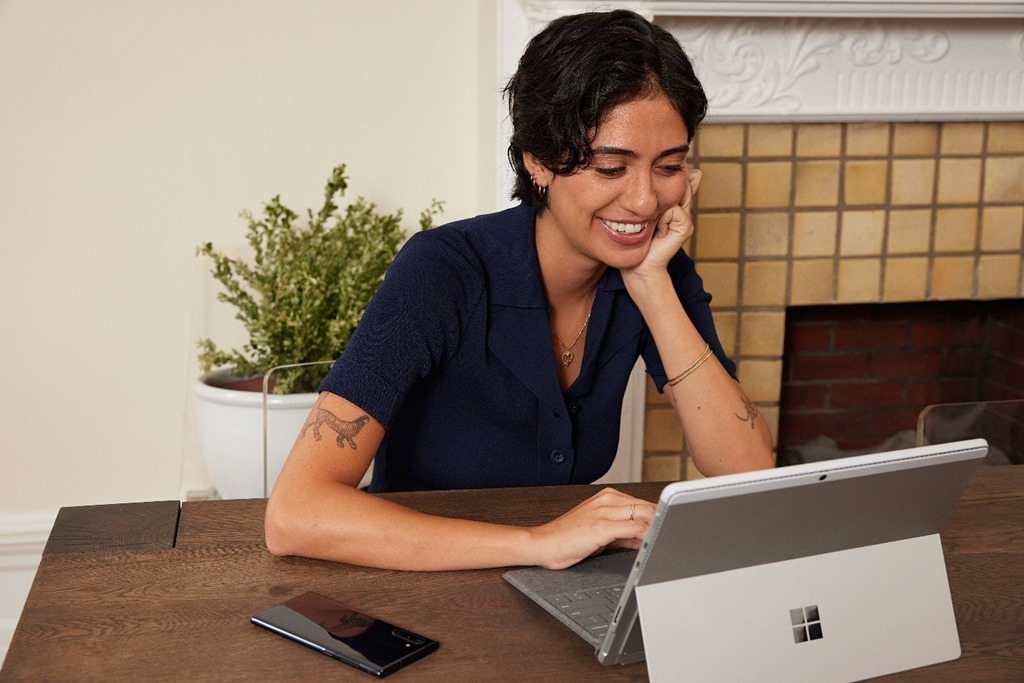 A woman sits at a table with a Microsoft tablet and a cell phone. She smiles at the tablet.